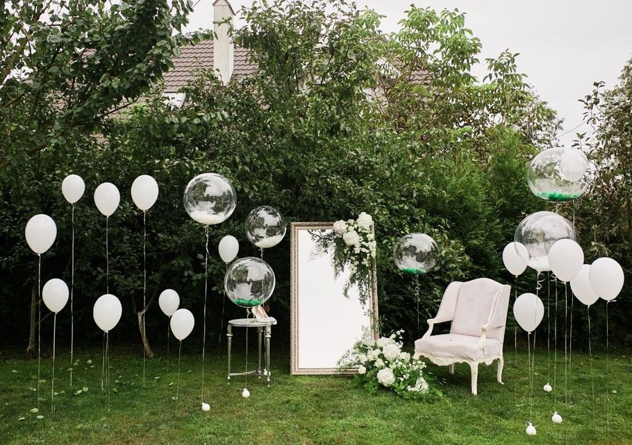 White balloon-decorated outdoor lounge with a chair, mirror panels, and lush green trees and grass