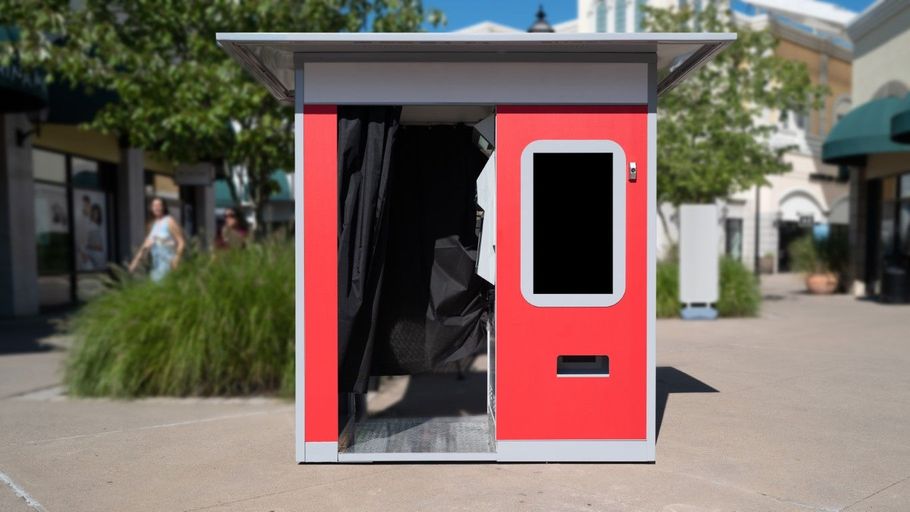 Small red kiosk or booth with an open door and window, standing outdoors on a sunny plaza.