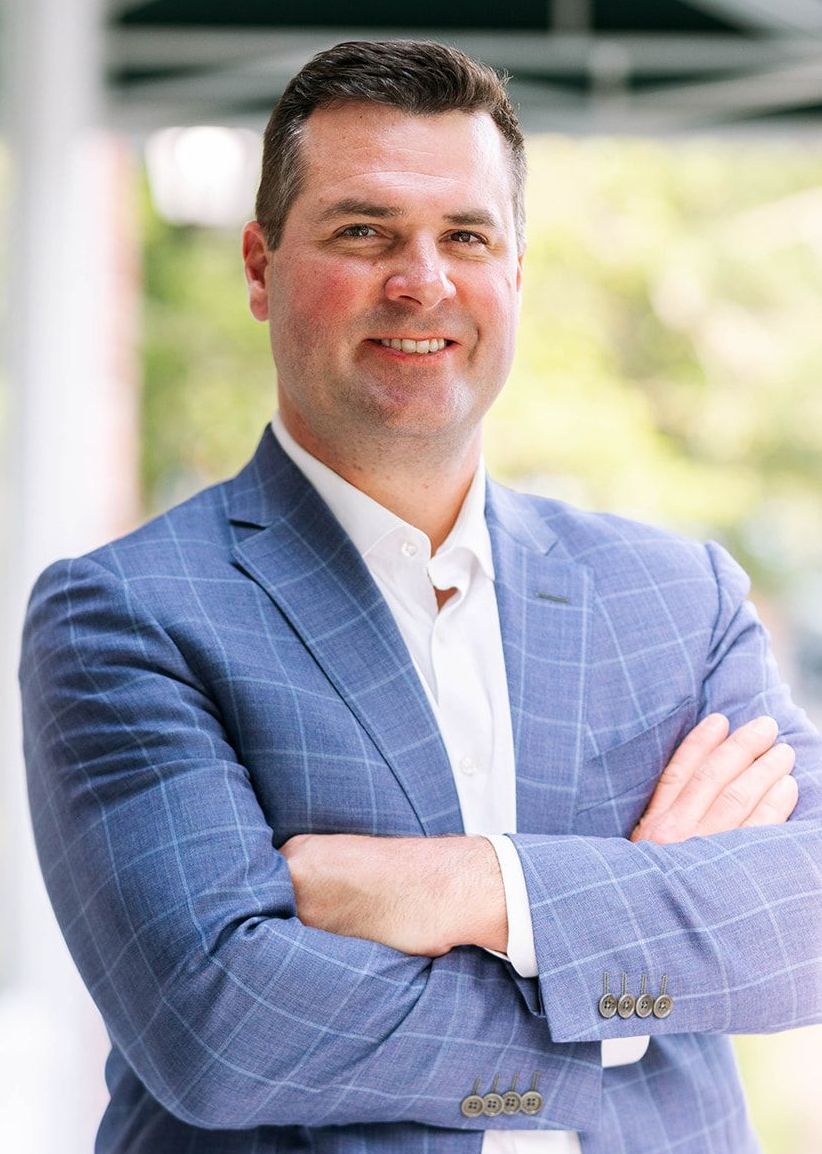 A man in a blue suit and white shirt is standing with his arms crossed and smiling.