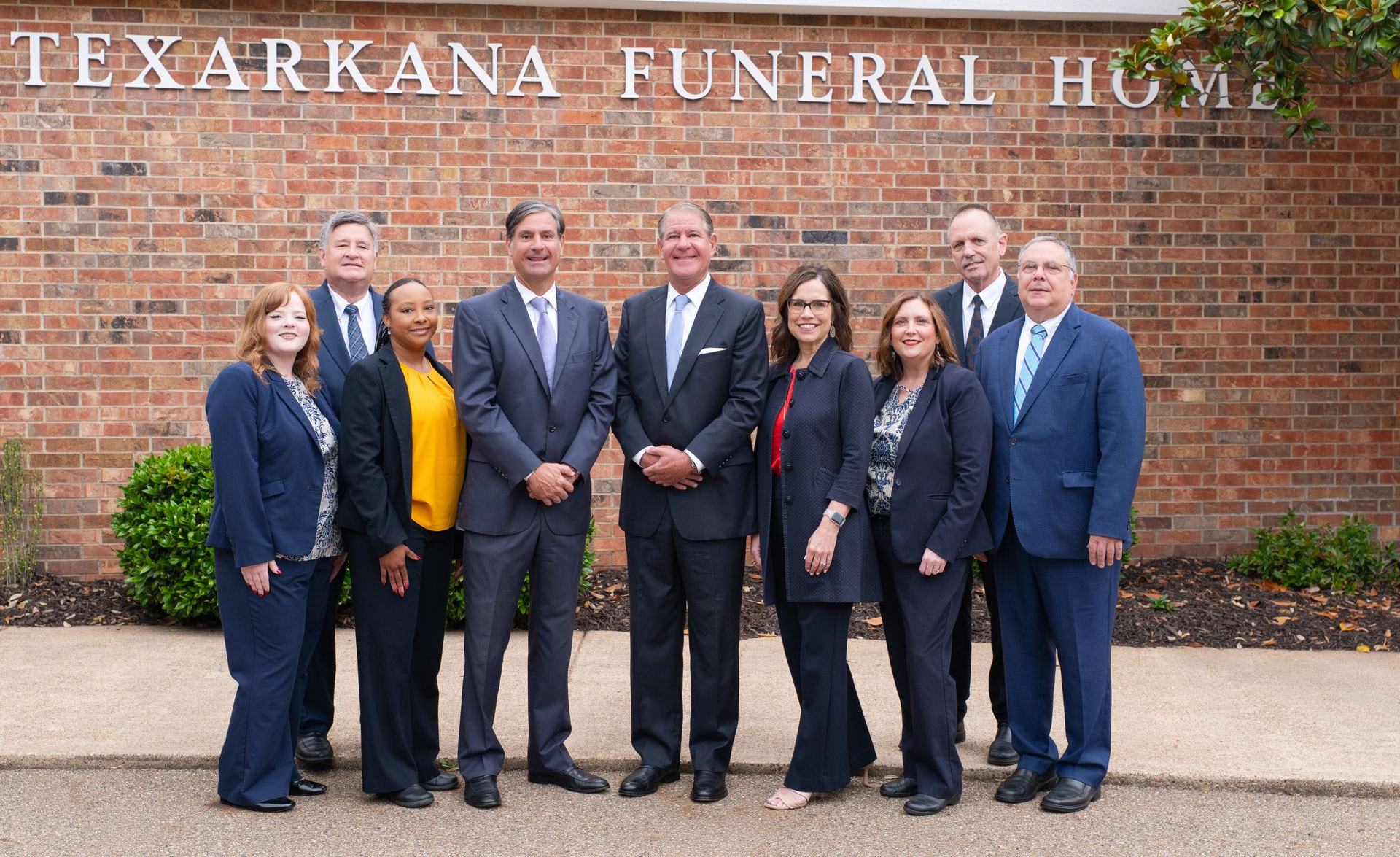 Eight people in business attire pose in front of a brick building labeled Texarkana Funeral Home.