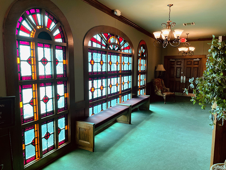 Bright hallway with stained-glass windows, green carpet, wooden bench, and chandelier lighting