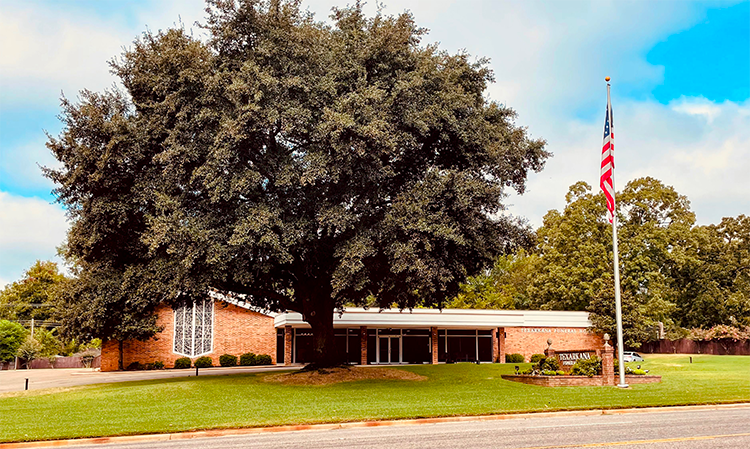 Low brick building with a large tree, green lawn, and an American flag on the right
