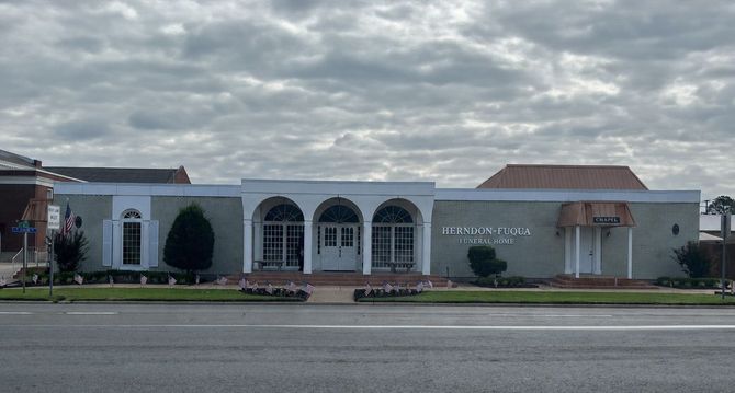 Row of storefronts under a cloudy sky, including a white building with arched windows and a small awning.