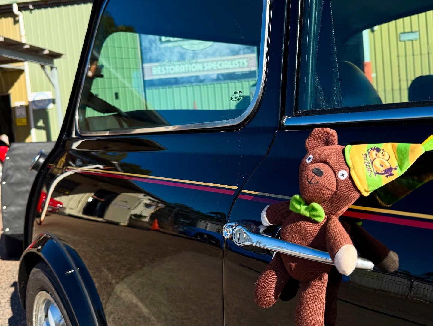 Black Mini Cooper with a teddy bear attached to the door handle, outdoors.