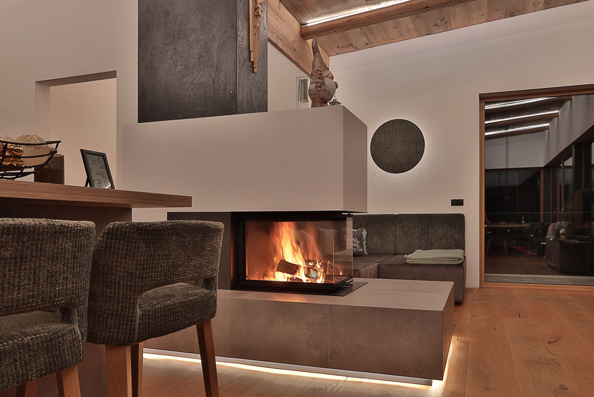 Modernes Wohnzimmer mit beleuchtetem Kamin, weißen Wänden, Holzelementen und geflochtenen Barhockern.
