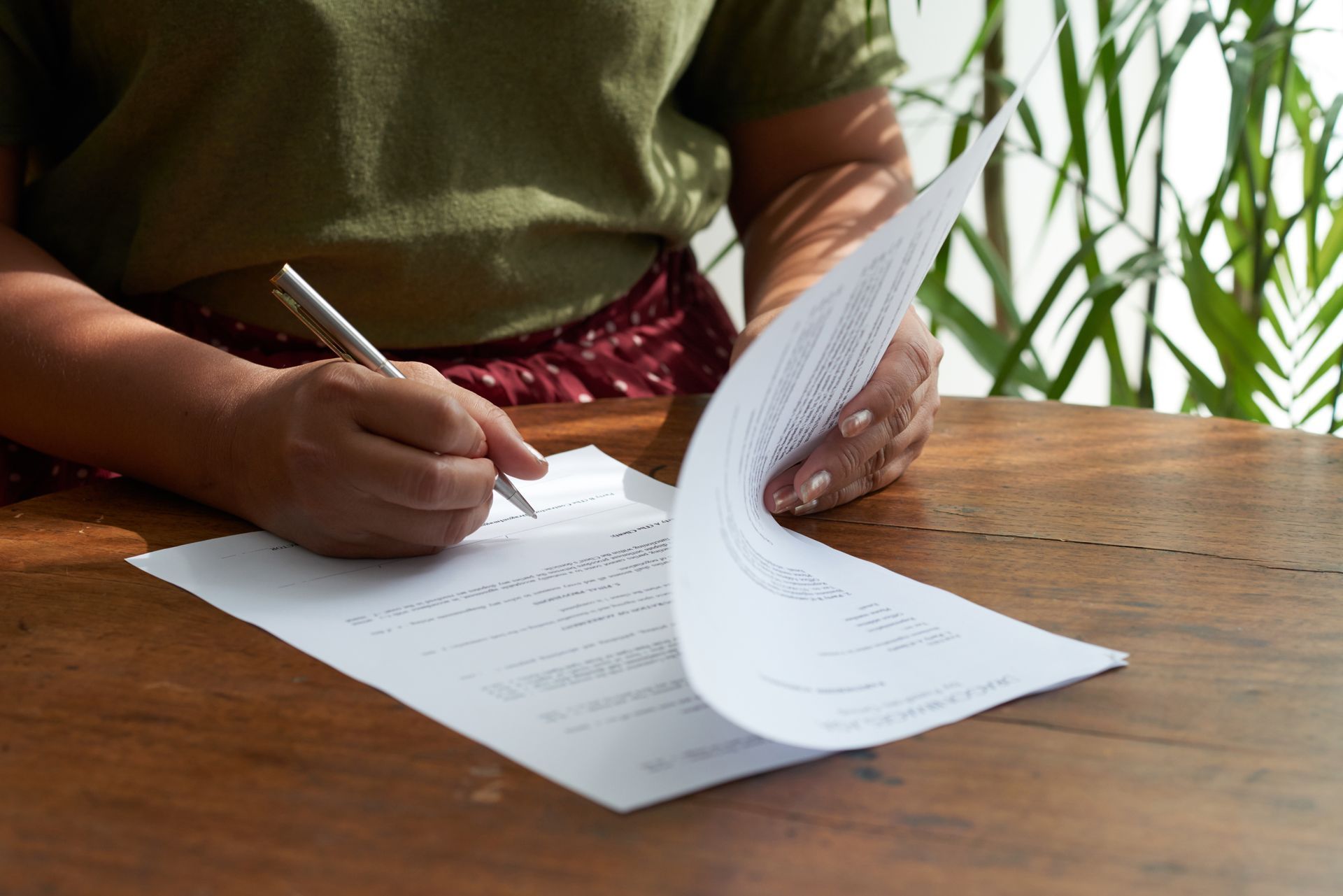 Person writing on paperwork at a wooden table, turning a page in a bright indoor setting.