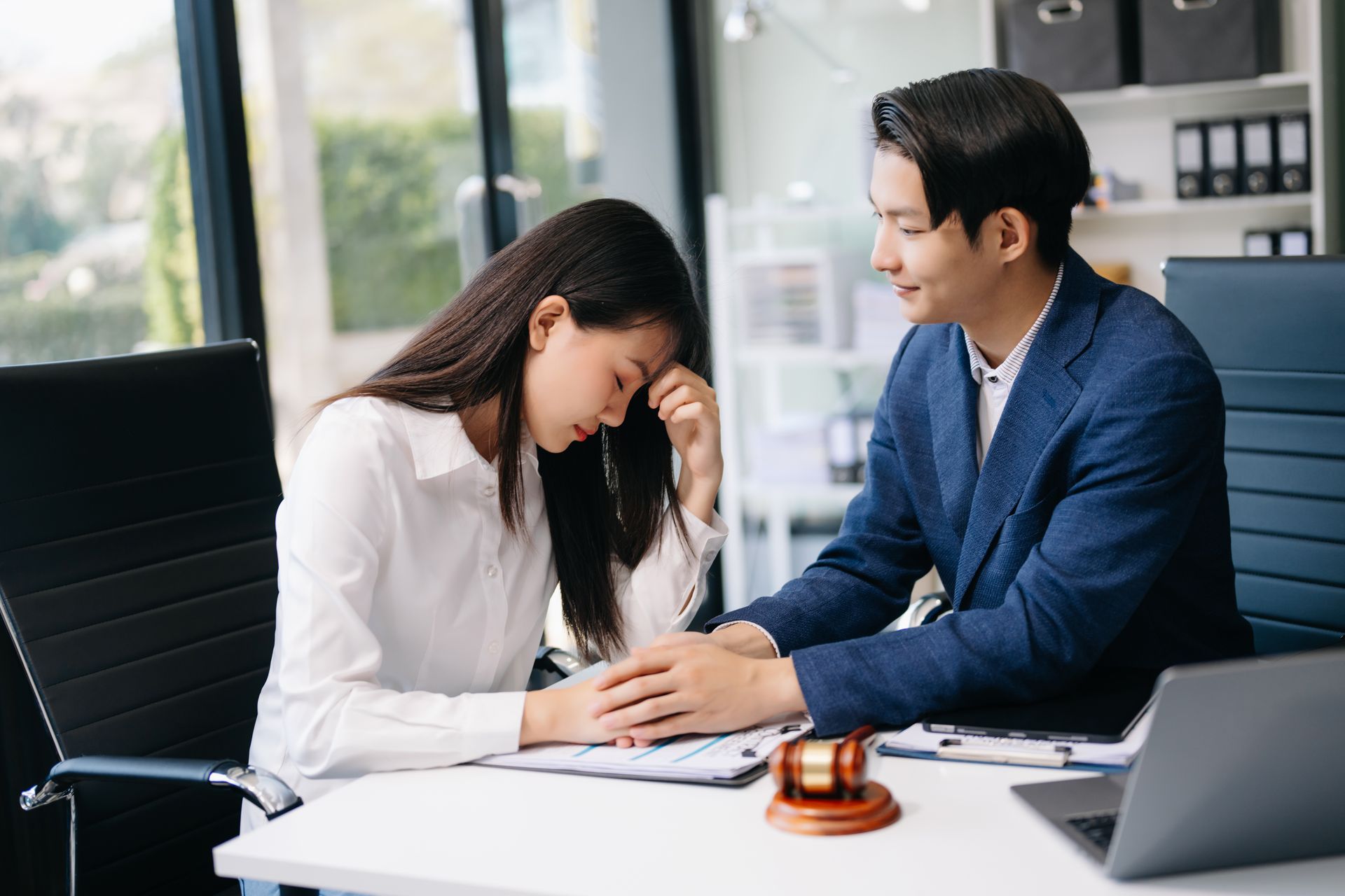 Two people sit at a desk in an office, one comforting the other as they hold hands.