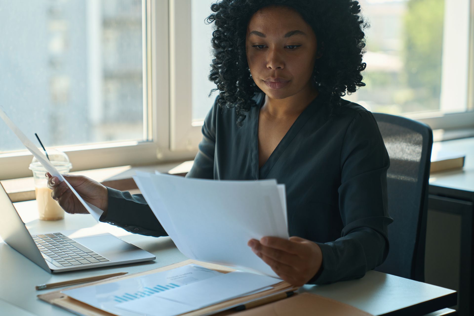 Woman reviewing documents at a desk beside a laptop in a bright office