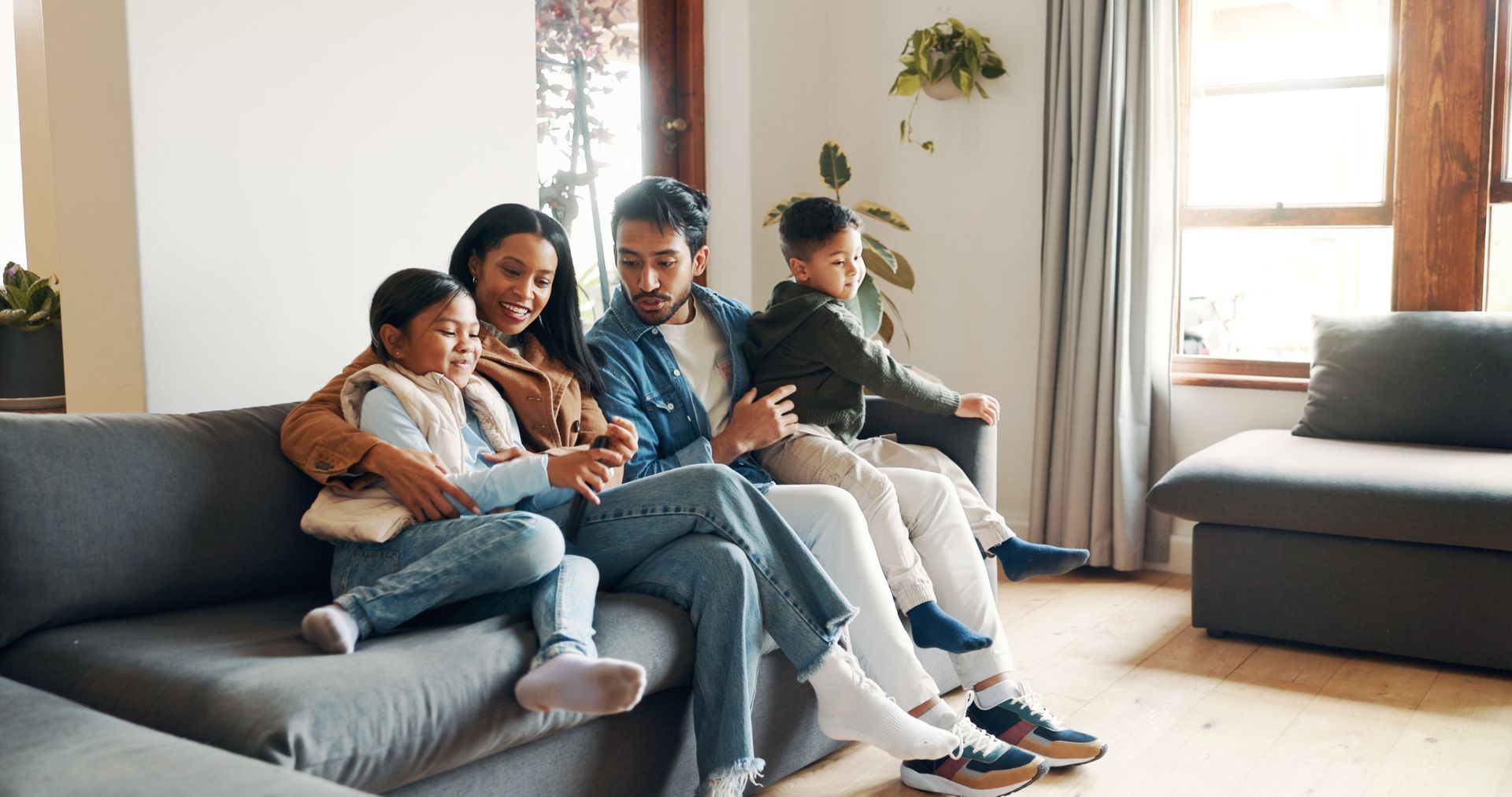 Family sitting together on a gray sofa in a bright living room, using a tablet and phone.
