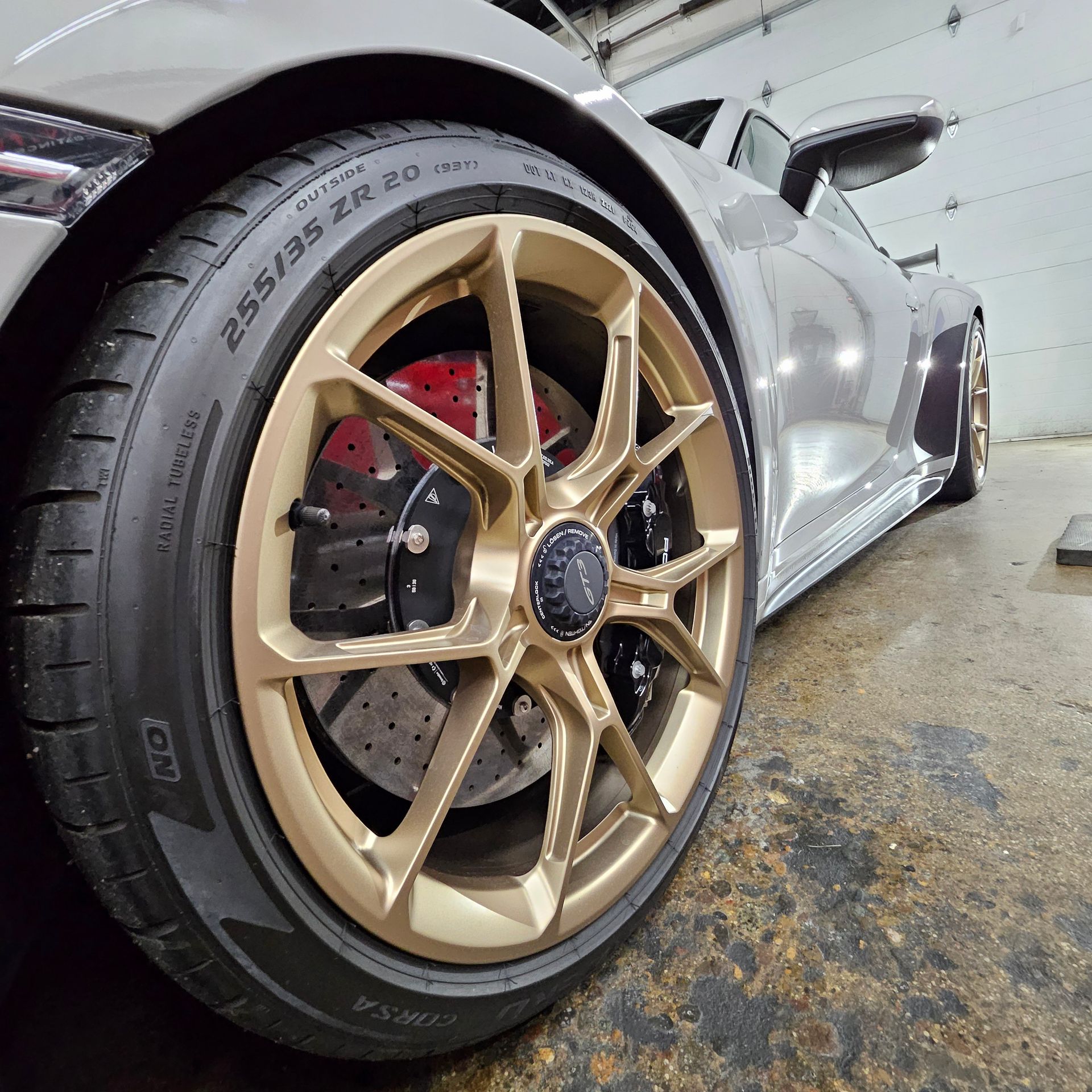 Close-up of a silver sports car’s gold alloy wheel and tire in a garage.