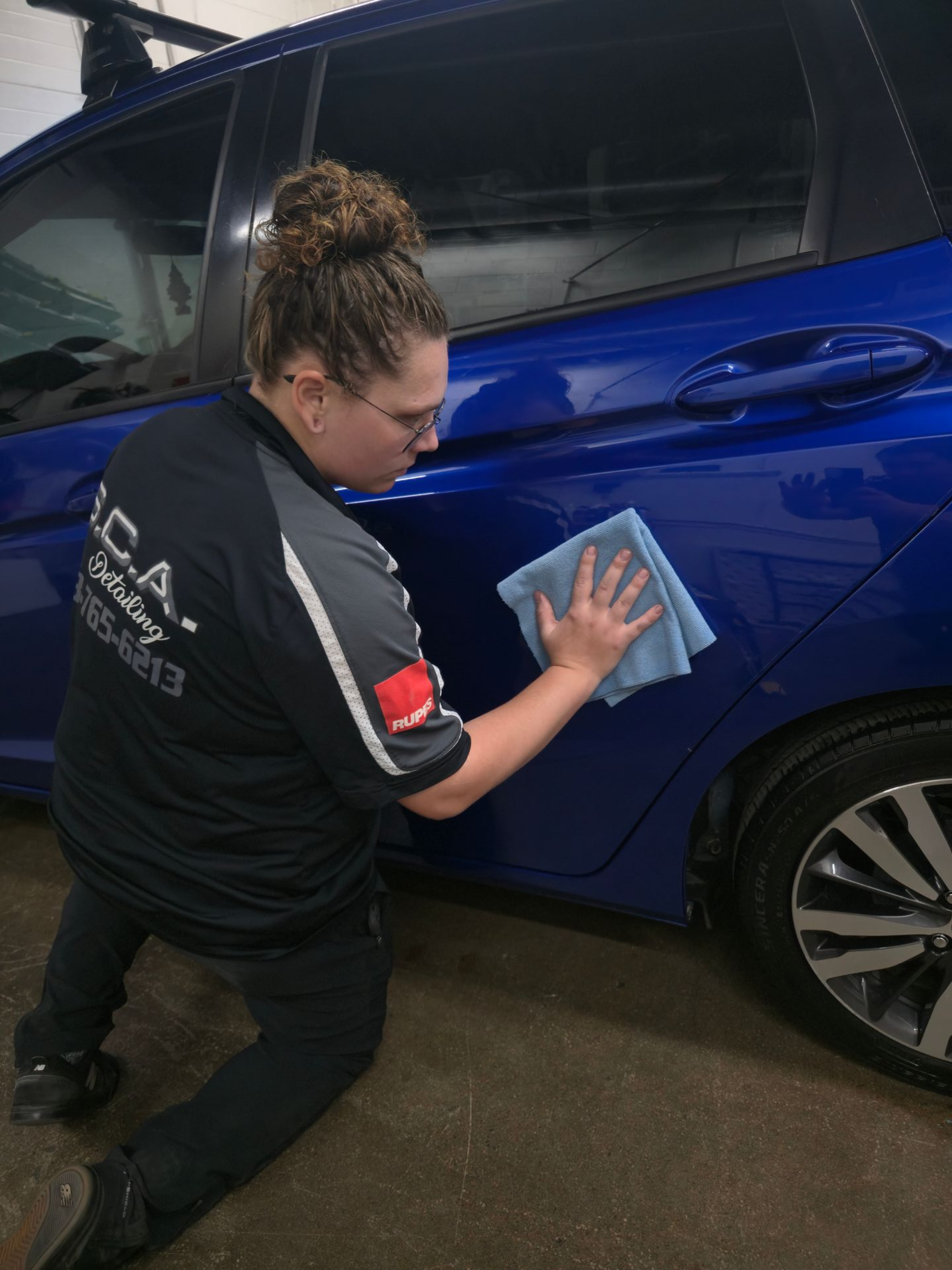 Person wipes a blue car’s side panel with a cloth in an indoor garage.