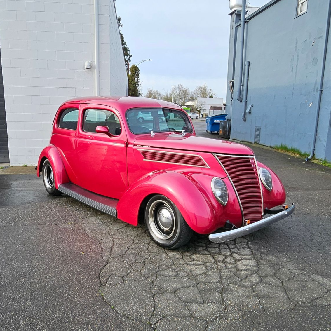Bright red vintage coupe parked on cracked pavement beside gray buildings