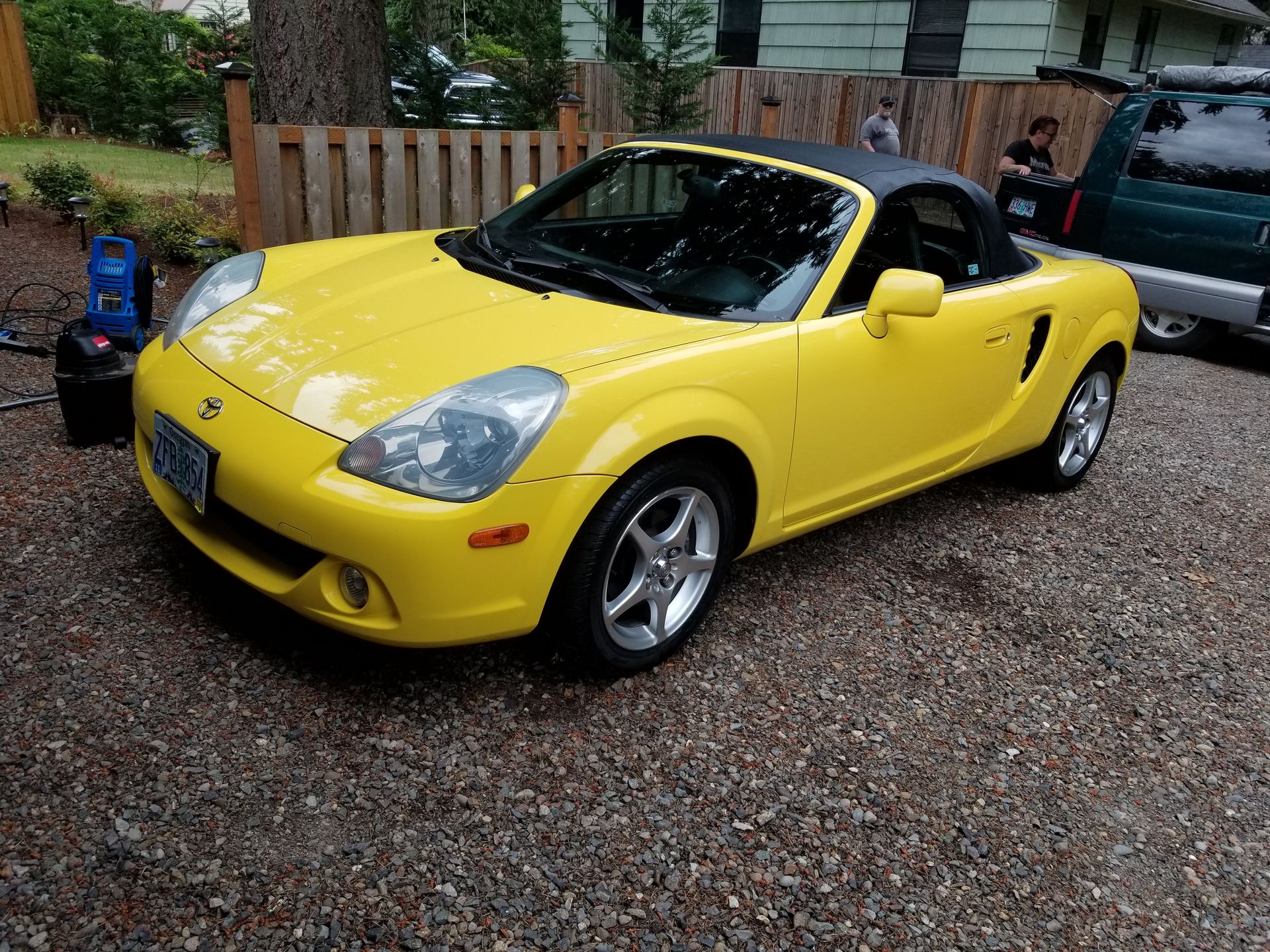 Yellow convertible sports car parked on a gravel driveway beside a wooden fence