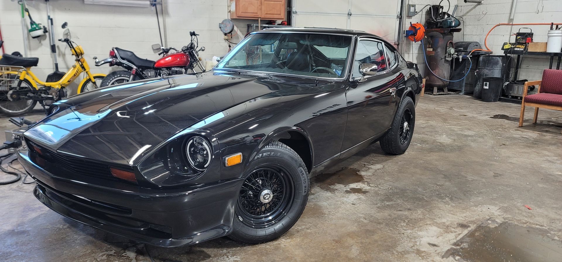 Black sports car parked in a cluttered garage, with tools and motorcycles in the background