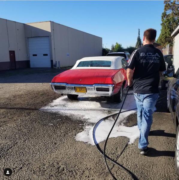 Man washing a red convertible car outside a building. White foam and water on the ground.