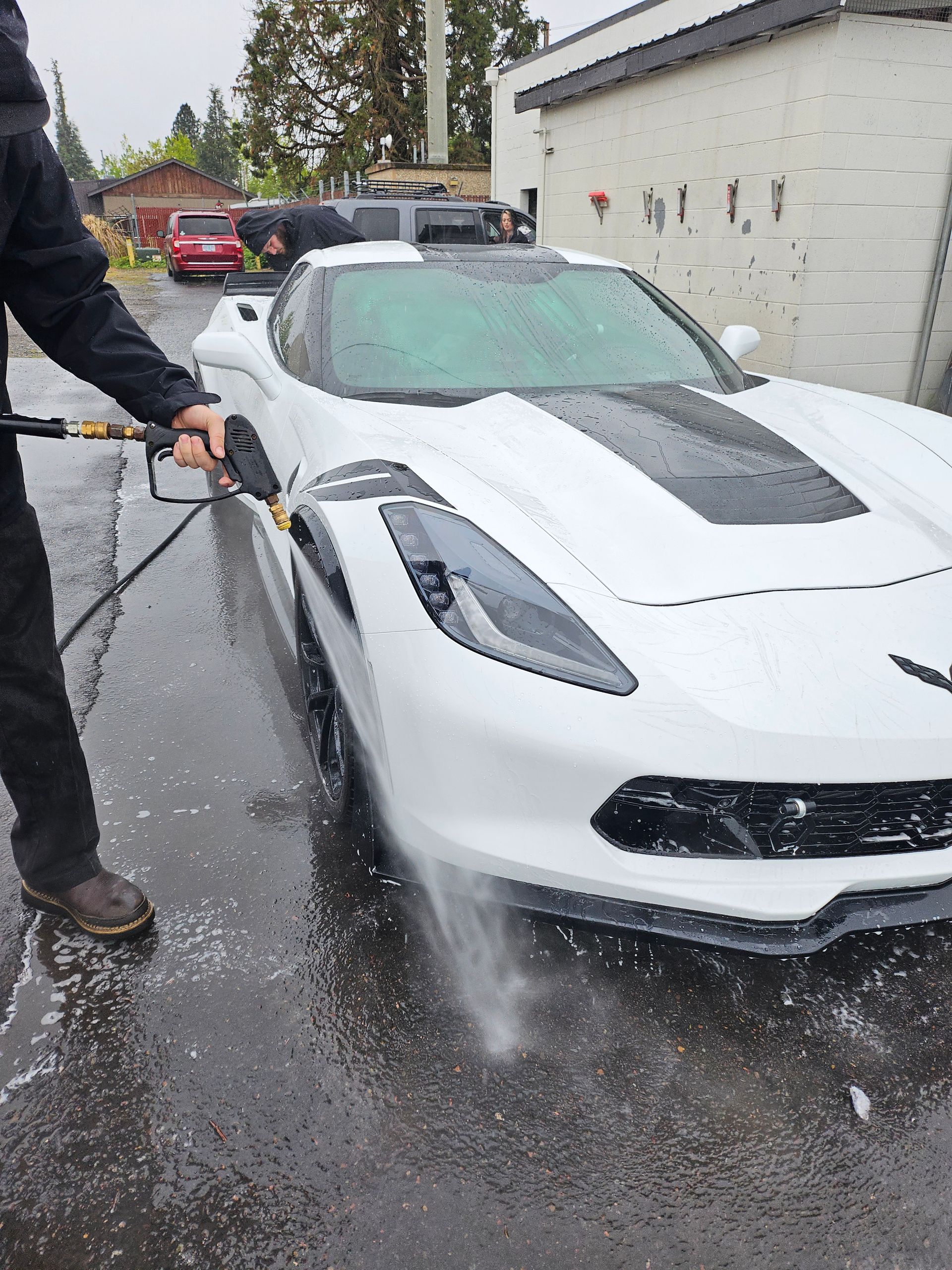 Person pressure-washing a white sports car in a wet driveway