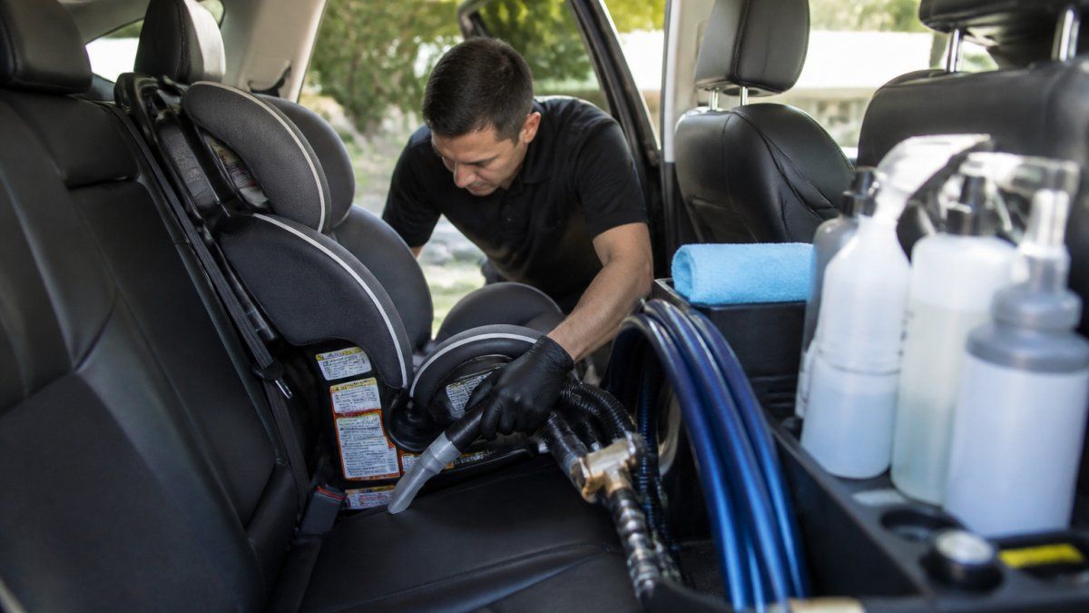 Professional detailer carefully cleaning and sanitizing around a properly installed child car seat in vehicle interior, with spot-free water system and cleaning supplies visible on work tray