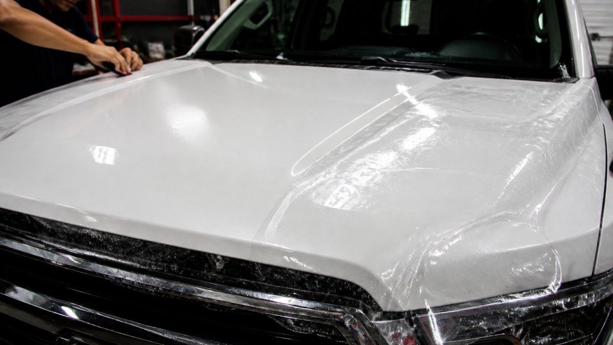 A close-up of a white pickup truck hood with clear paint protection film partially applied, showing the gloss difference between protected and unprotected areas