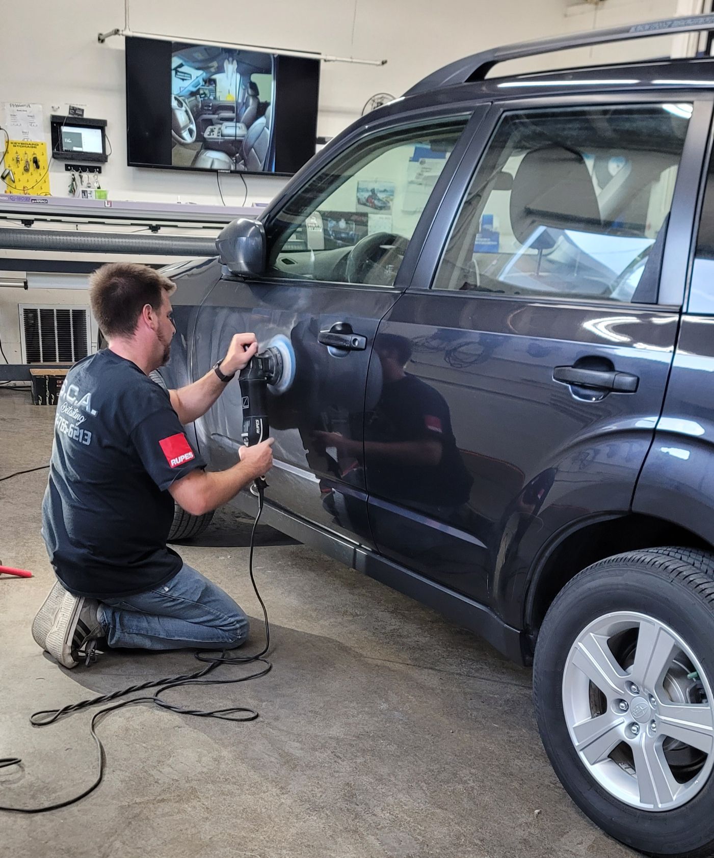 Technician repairing a dark SUV’s door in a garage, using a power tool while kneeling on the floor.