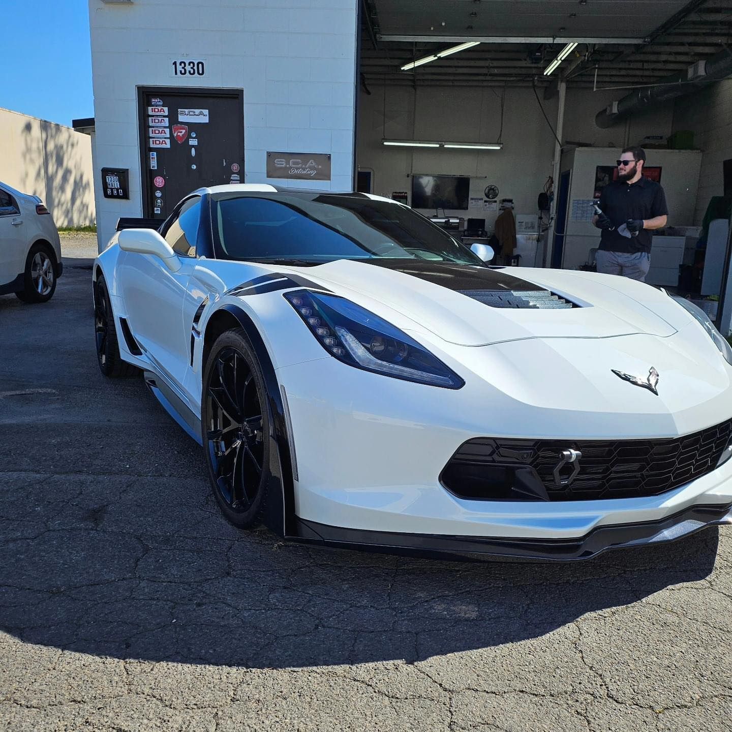 White Chevrolet Corvette parked outside a garage, with a man standing near the open bay.