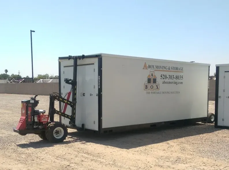 White storage trailer in a sandy lot, with a red bike-like machine beside it under a clear sky.