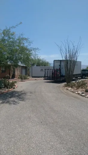 Gravel driveway leading to a white trailer and parked RV beside sparse desert trees under a clear sky