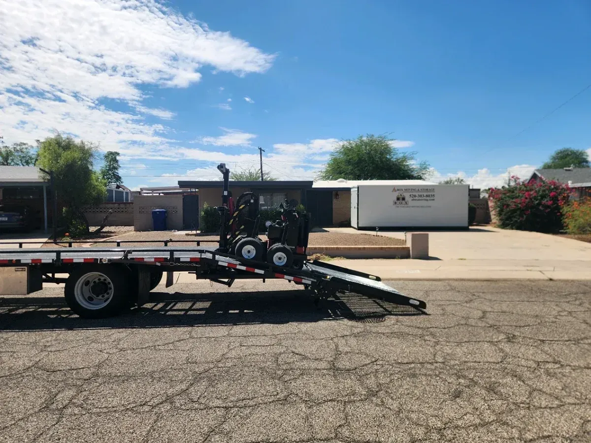 Flatbed trailer with a ramp parked on a cracked lot under a blue sky, with buildings and a white container in back