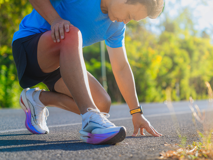 Runner crouching on a road, touching his knee while tying a white shoe in bright sunlight.