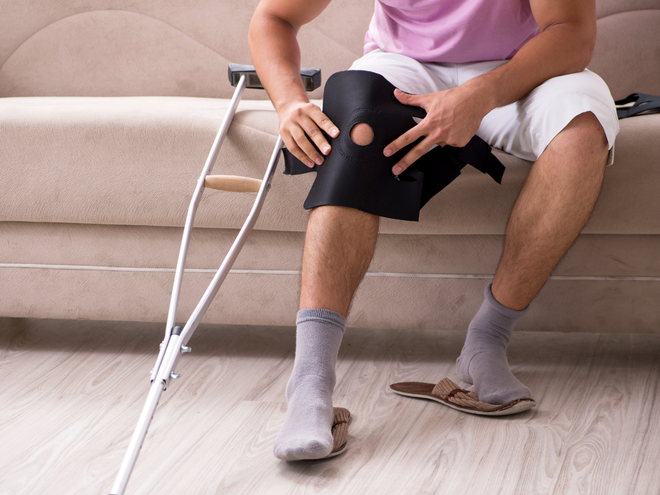 Person seated on couch with knee brace and crutches, adjusting injured leg in a living room