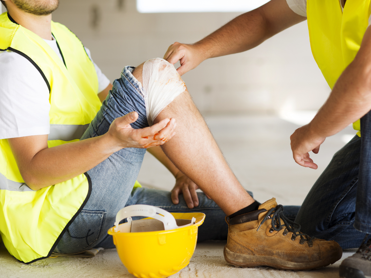 Two workers in yellow safety vests applying a bandage to a knee beside a hard hat on the floor