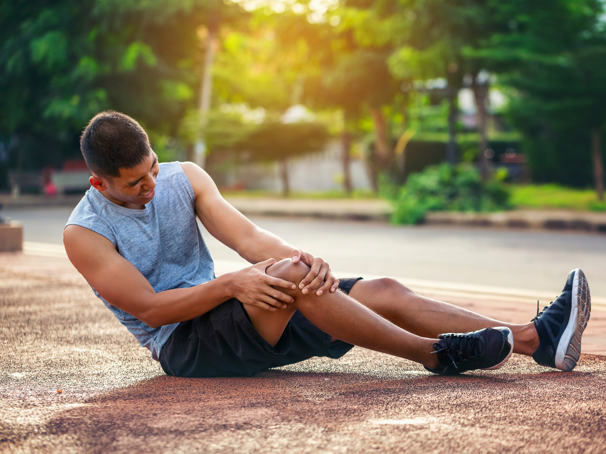 Person stretching leg outdoors on a sunlit path, wearing a sleeveless top and shorts.