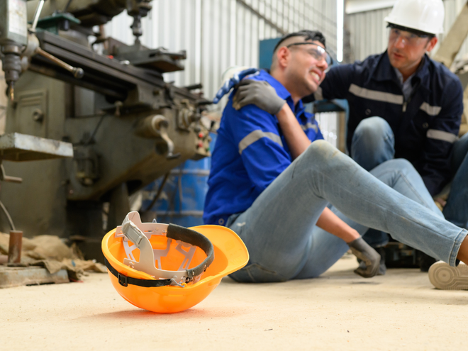 Worker sitting on floor near a hard hat, being assisted by a coworker at an industrial site
