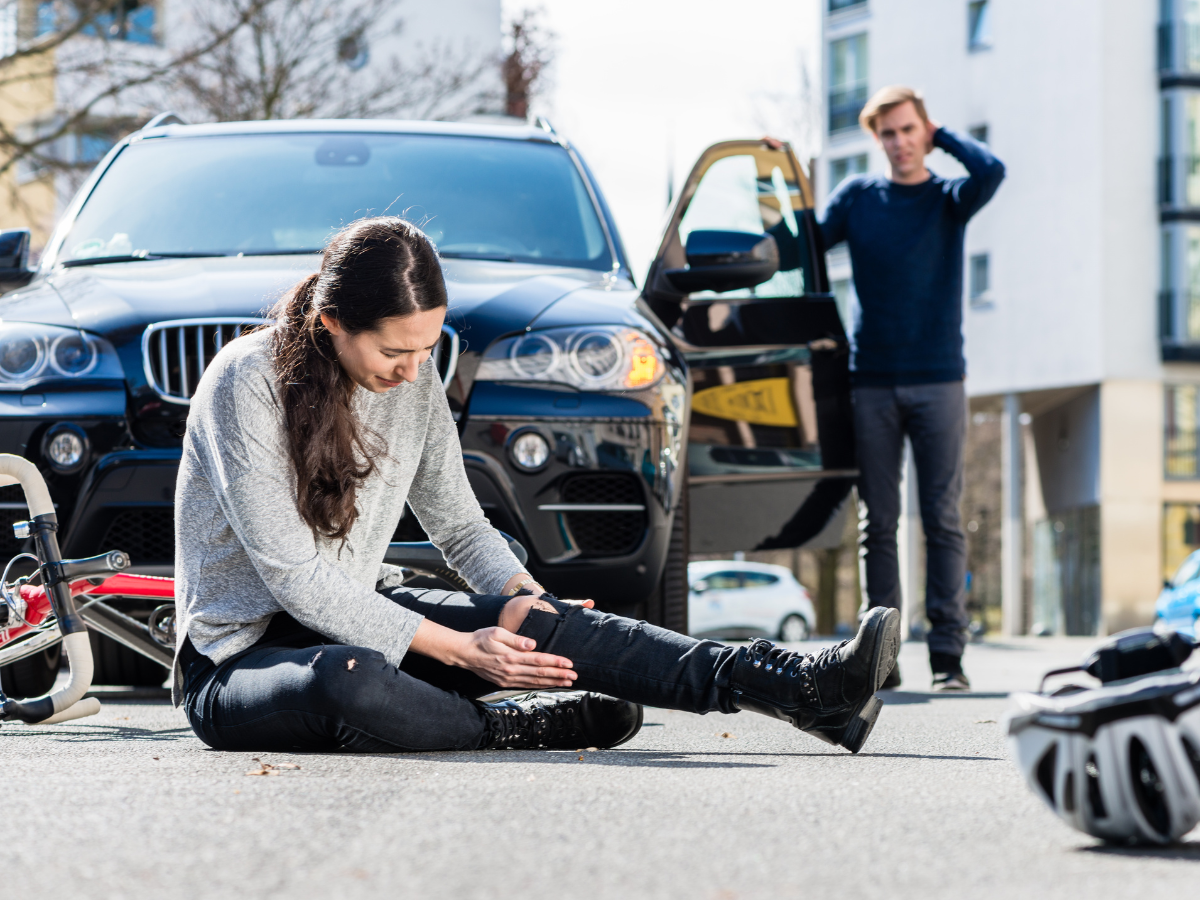 Woman sits on pavement holding injured leg beside SUV while a man stands nearby, looking concerned