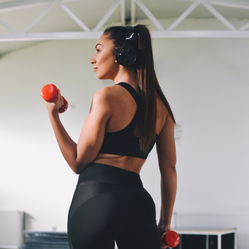 Woman in workout clothes lifting red dumbbells in a gym, seen from behind.