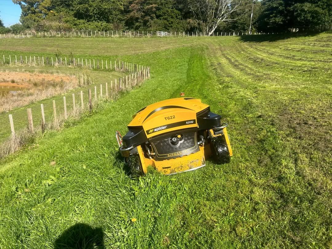 Yellow robotic lawnmower on a grassy hillside, cutting a strip of grass. Fence and trees in the background.