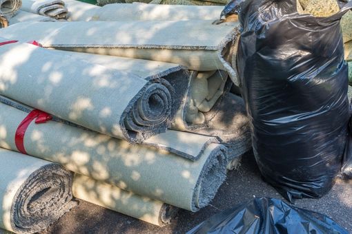 Rolled carpets or mats stacked outdoors beside a black trash bag, in dappled sunlight