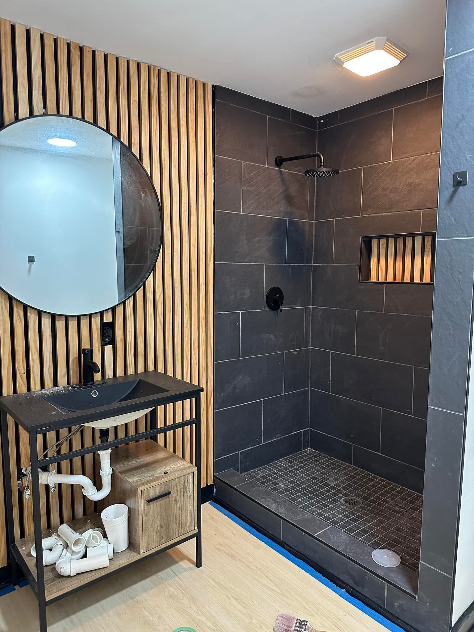 Modern bathroom: black shower, wood accent wall, circular mirror, black sink.