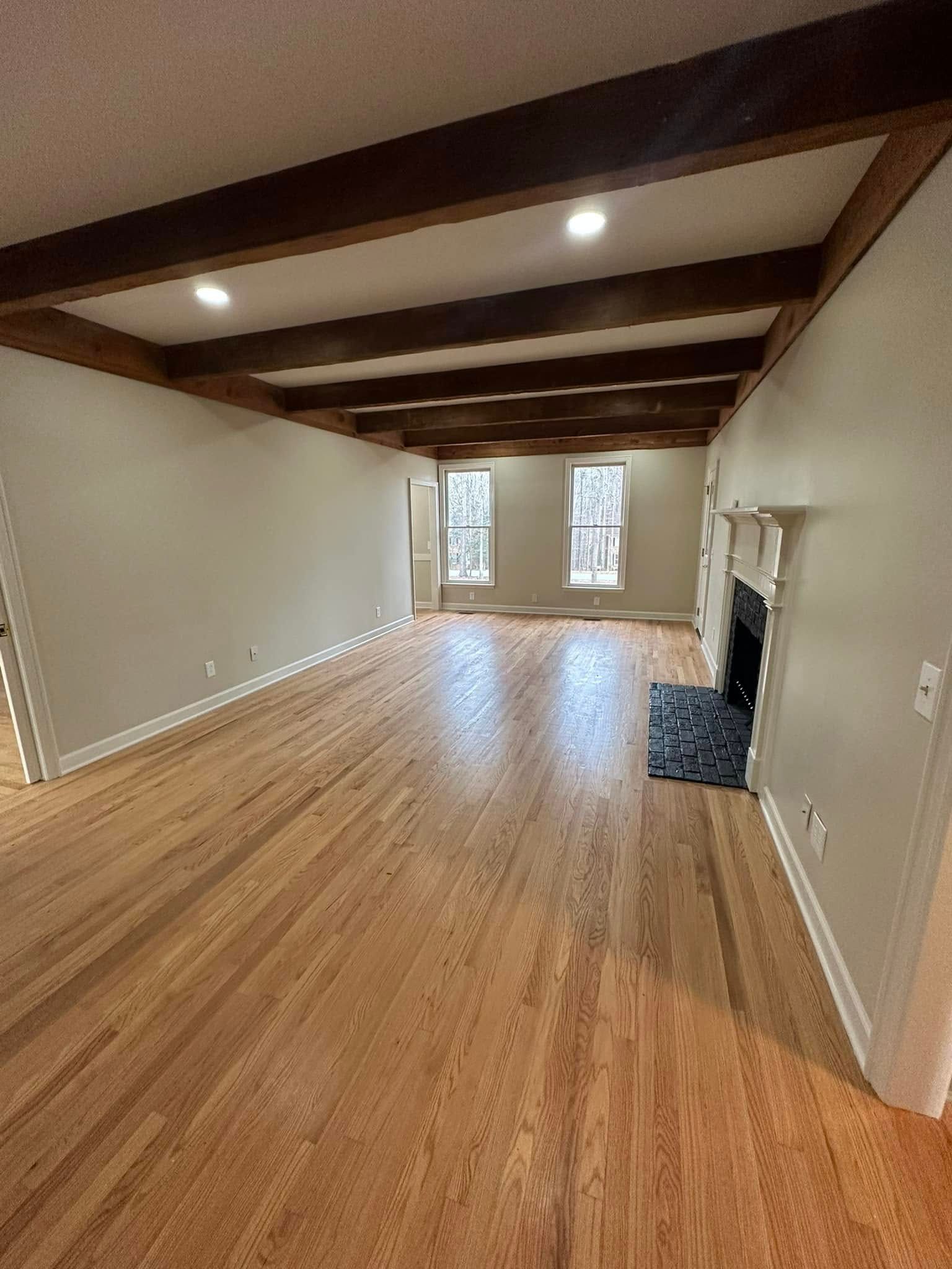 Spacious living room with wood flooring, ceiling beams, fireplace, and two windows.