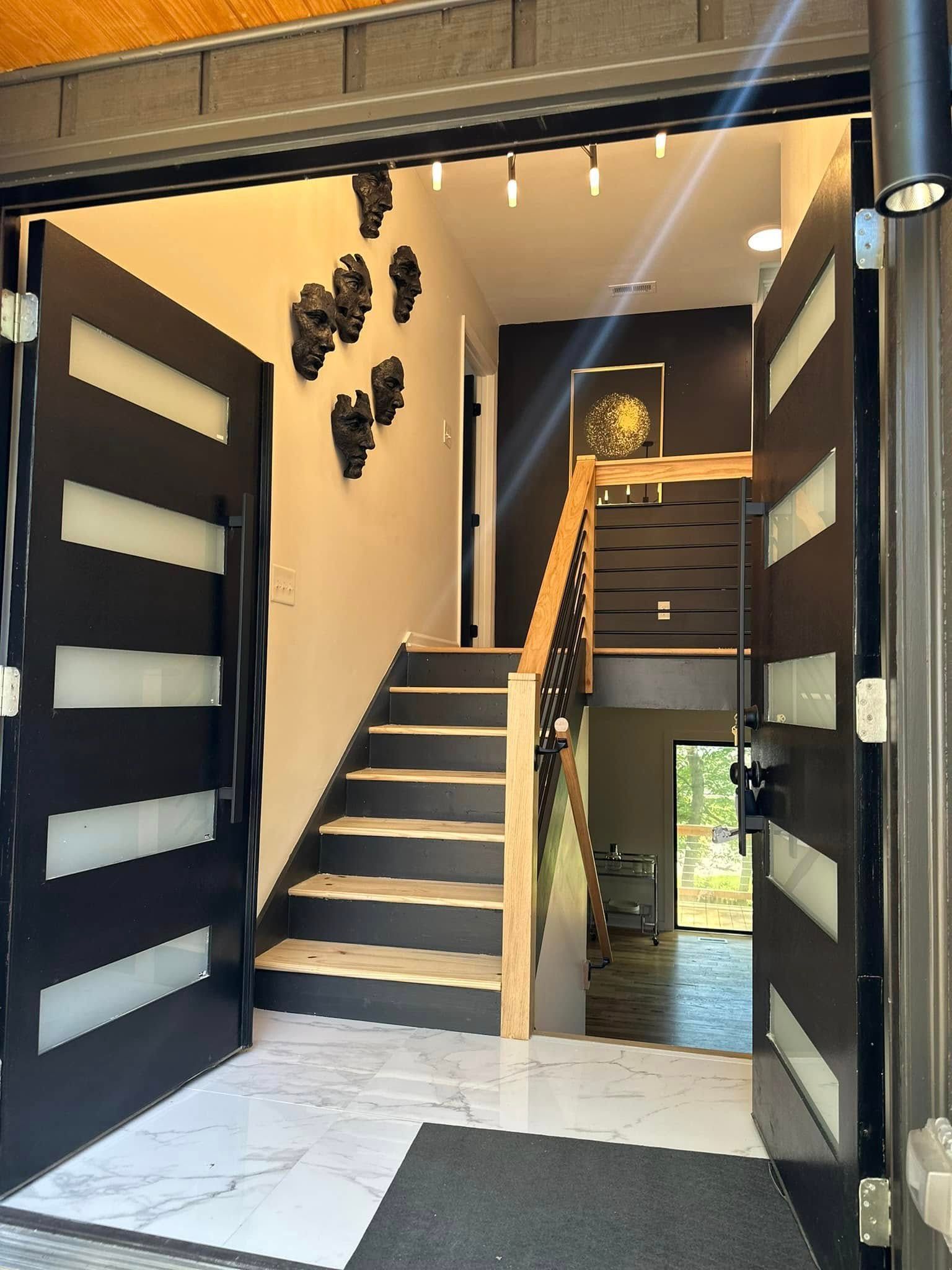 Open entryway with double black doors, leading to stairs. White marble floor, artwork on wall.