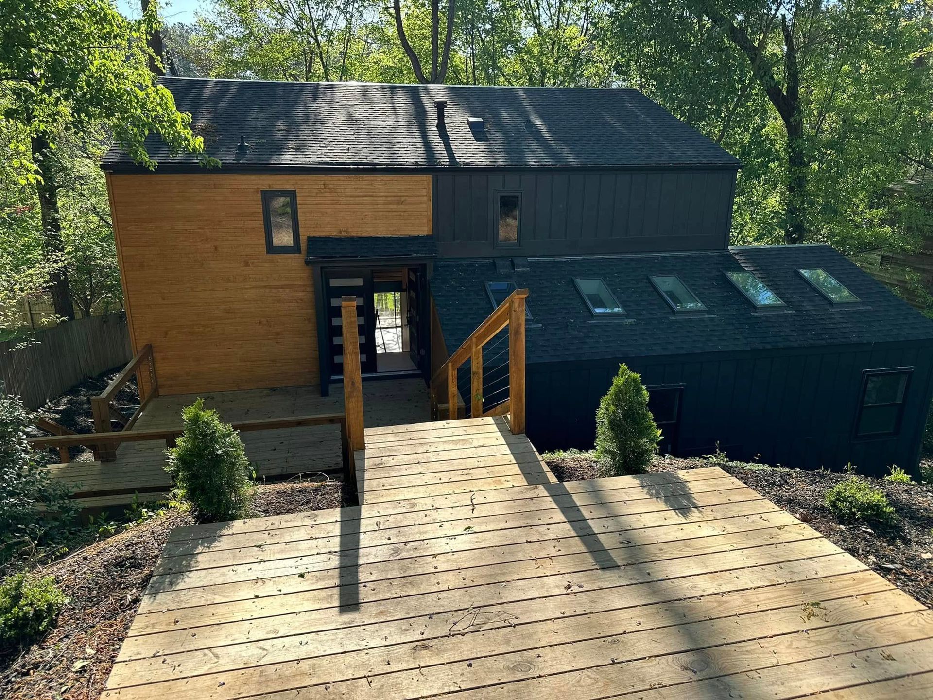 Wooden deck leads to two-toned house: light wood and dark grey exterior. Skylights and greenery surround.