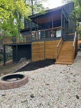 Cabin with dark exterior, wood deck, and fire pit in a wooded setting with gravel and mulch.