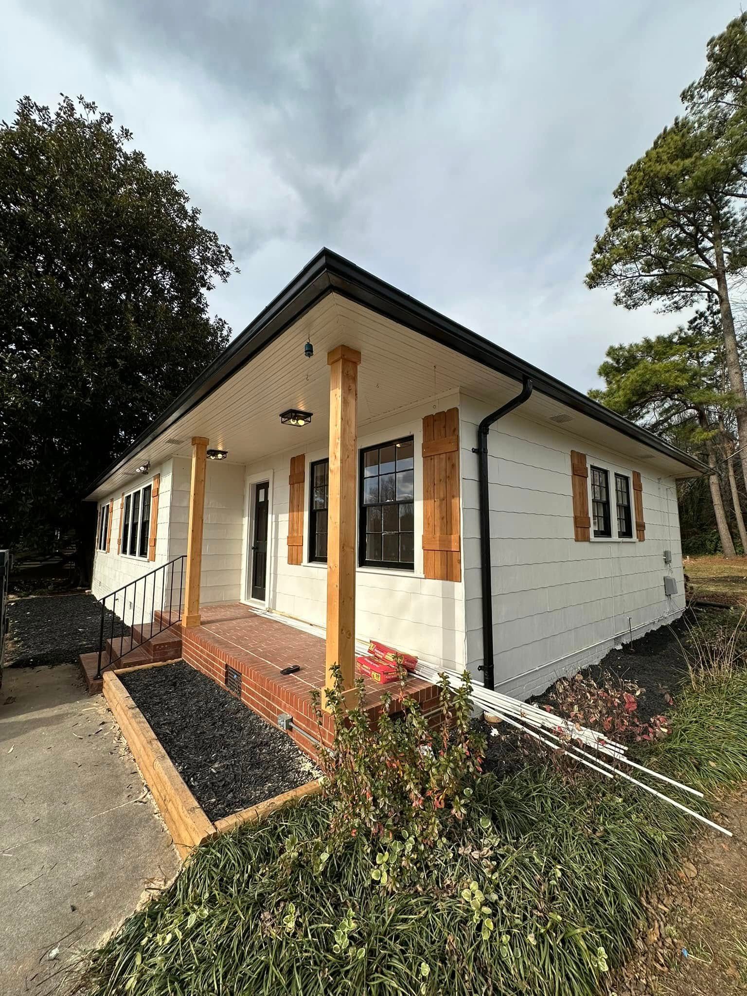 White house with brown shutters and porch. Black roof and trim. Trees and cloudy sky in background.