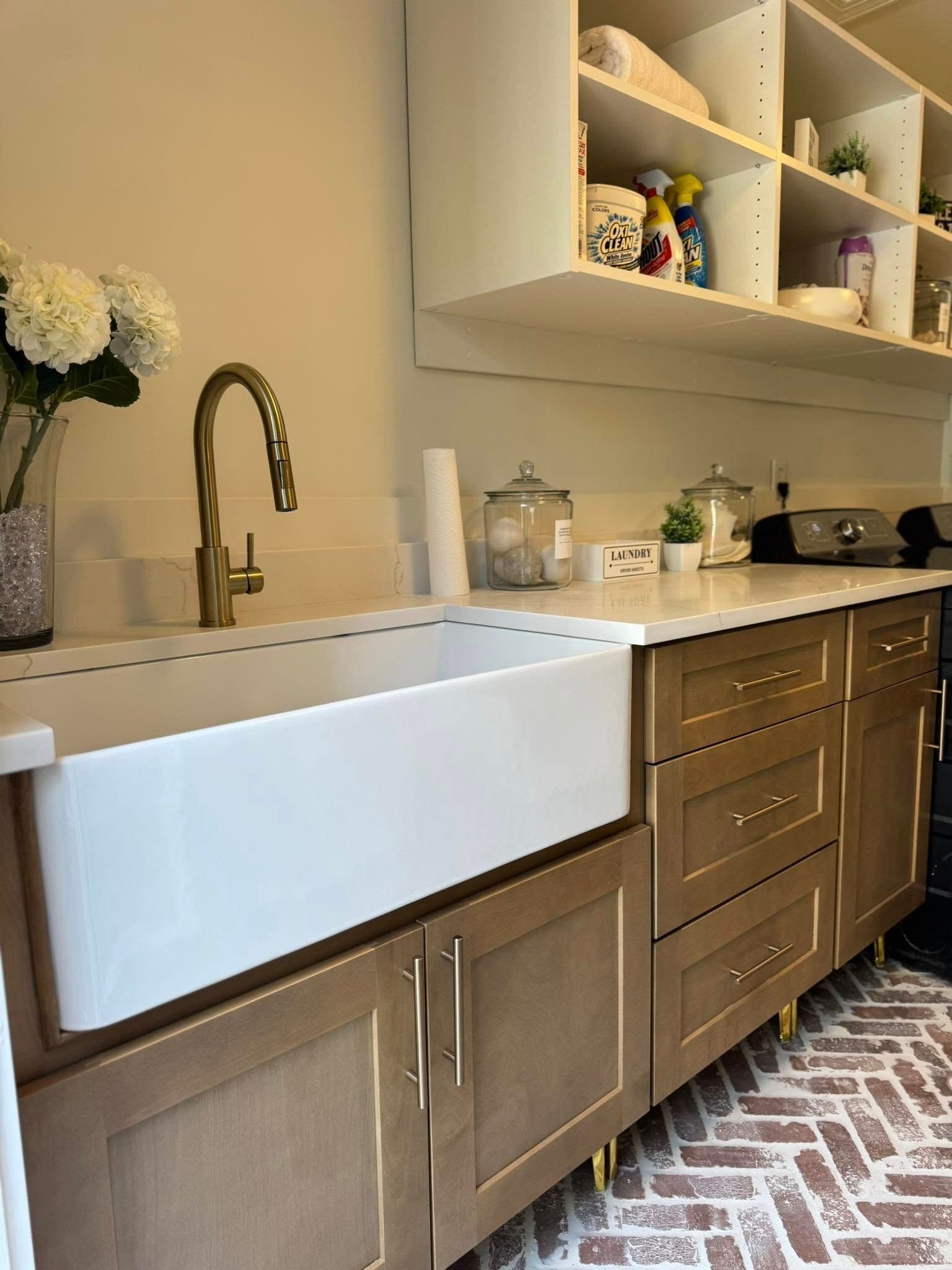 Laundry room with white sink, gold faucet, and cabinets. Brick floor, open shelving, and flowers.