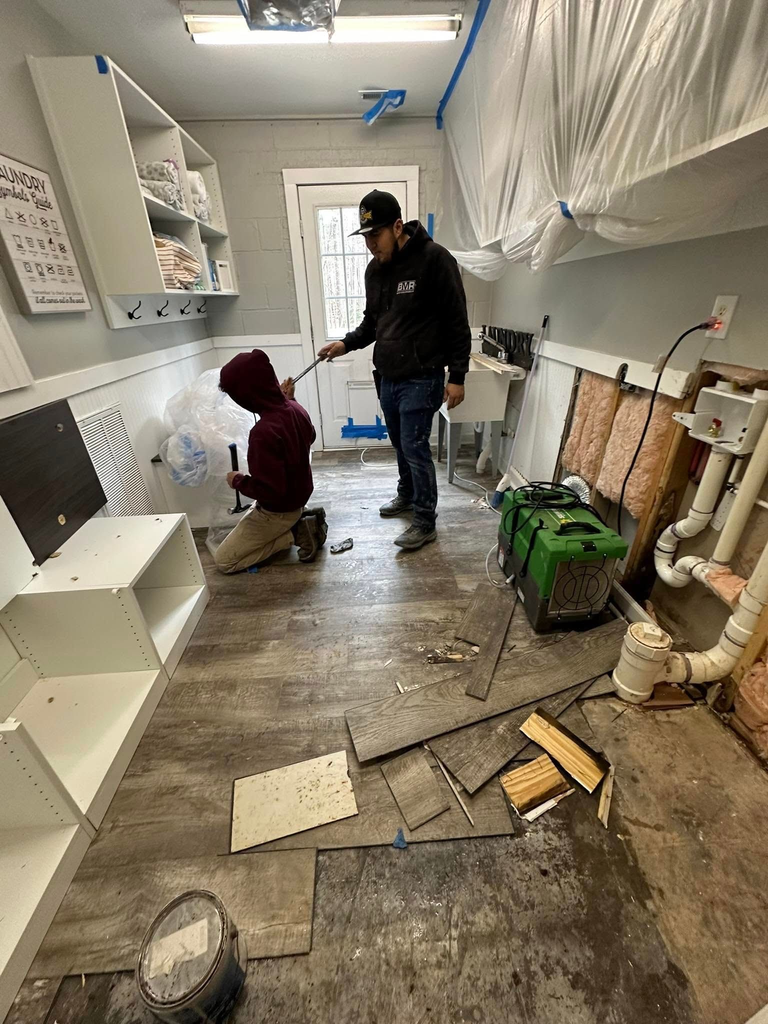Two people remodel a small room: one kneels, the other points. Construction materials and tools are scattered on the floor.