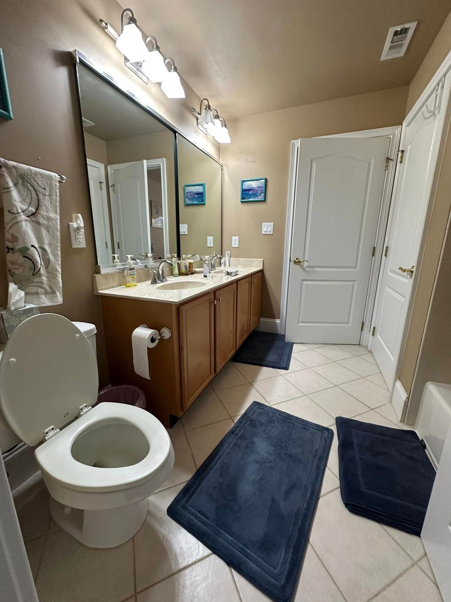 Bathroom with a toilet, vanity with double sinks, and two blue bath mats on the tiled floor.