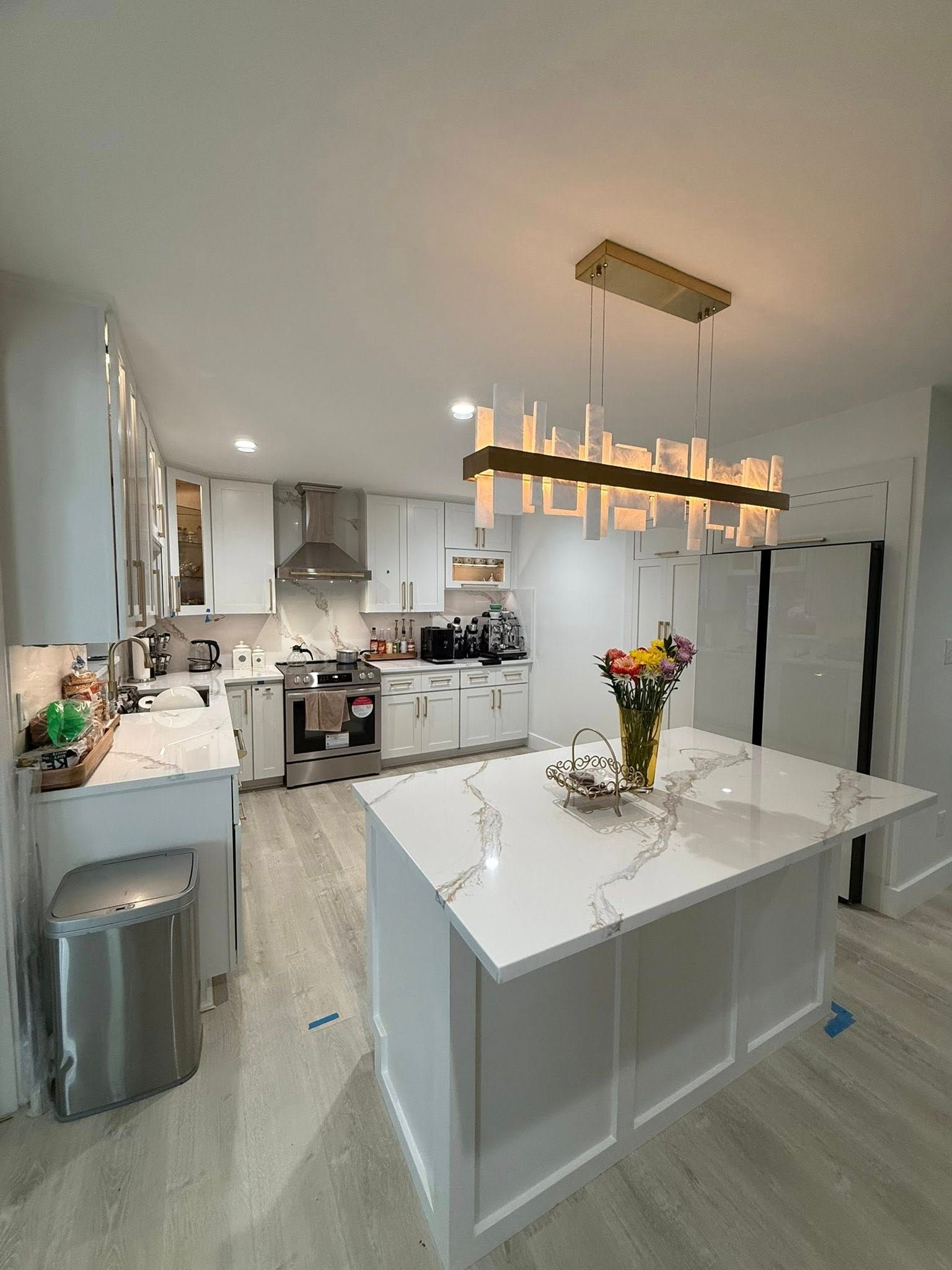Modern white kitchen with island and gold light fixture. Countertops are white with gray veining.