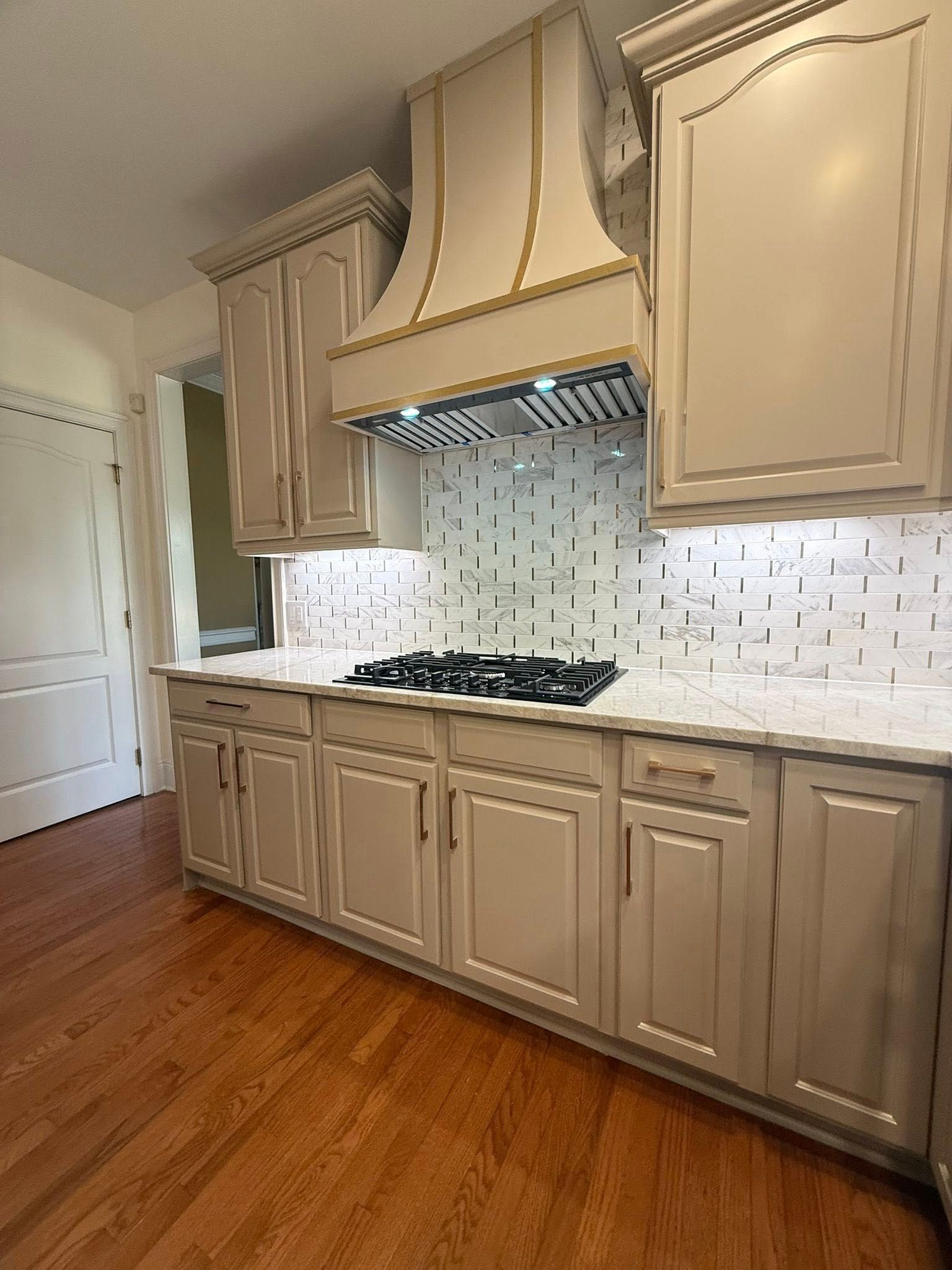 Beige kitchen with stove, range hood, cabinets, white backsplash, and wooden floor.