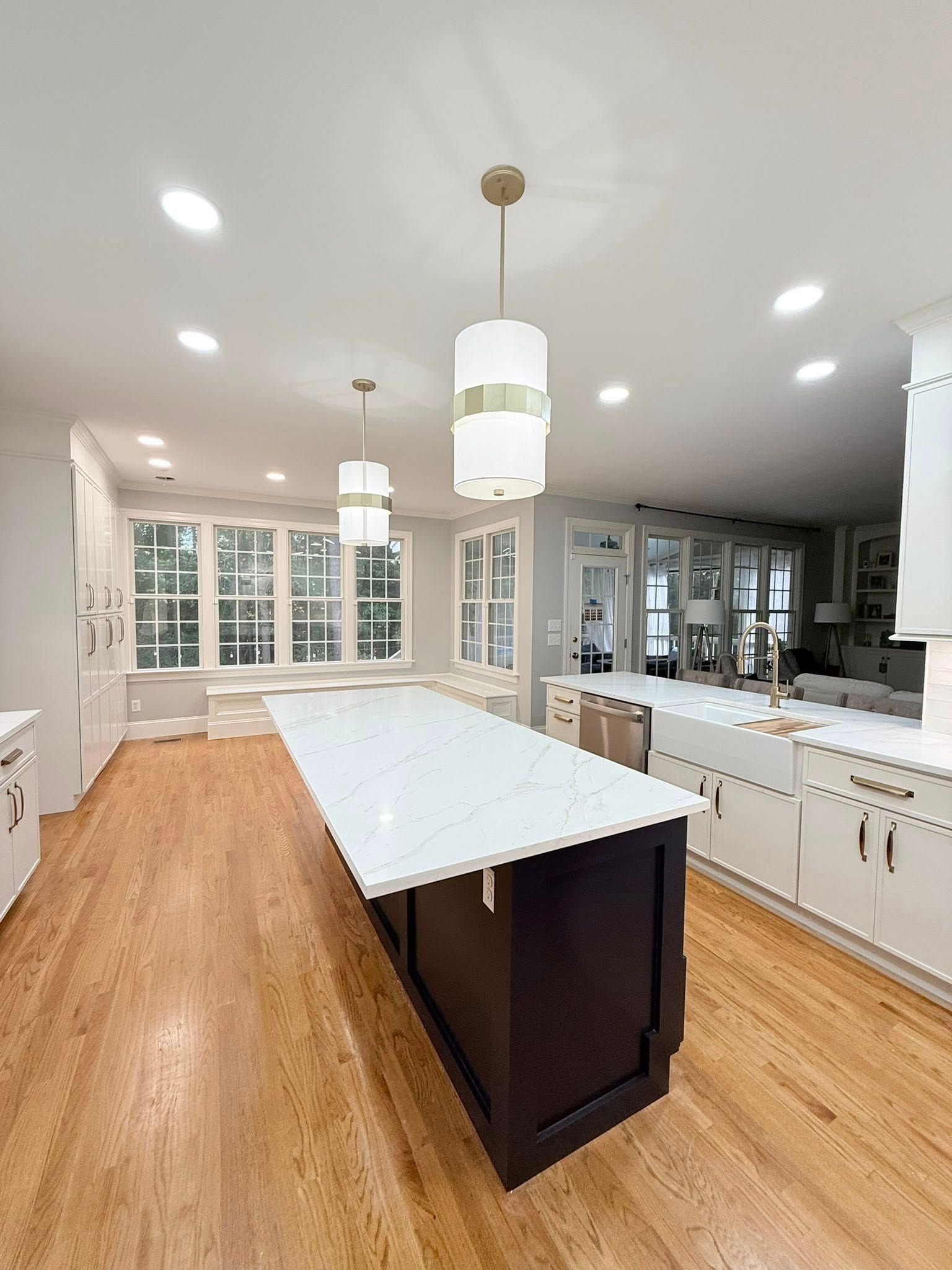 Modern kitchen with a dark island, light countertops, and pendant lights. Wooden floors and large windows.