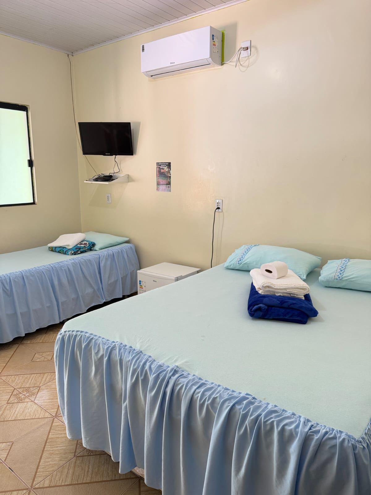 Um quarto de hotel com duas camas, colchas azul-claras, uma televisão na parede e um aparelho de ar condicionado acima.