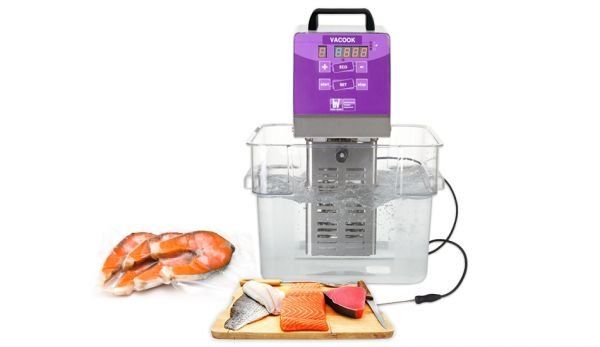 Un fornello sous-vide viola immerso in un bagno d'acqua trasparente, accanto a salmone e gamberi sigillati su un piatto.